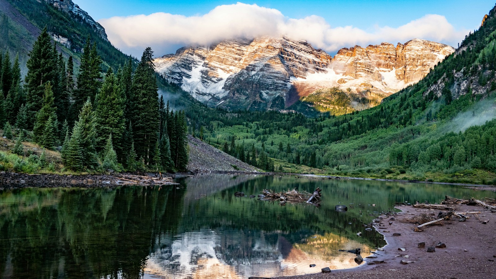 Mountains, evergreens, and a lake reflected in still water.